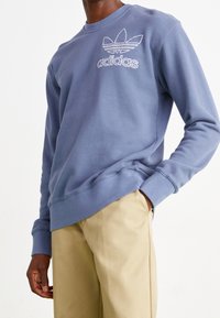 Modrá mikina z bavlny, s kulatým výstřihem, žebrovanými manžetami a vyšívaným logem Adidas na hrudi. V kombinaci s béžovými kalhotami.