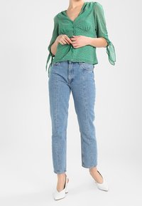 Blouse verte à pois avec col en V, taille froncée et manches nouées. Associée à un jean bleu clair taille haute à jambes droites et des mules blanches à talons.