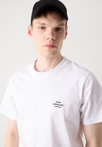 Hvid bomulds t-shirt med et lille sort broderet logo på venstre bryst. Rund hals, korte ærmer, glat tekstur.