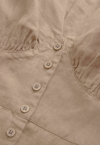 Haut en lin beige avec un corsage froncé, cinq boutons de fermeture avec quatre trous chacun, et des détails de couture subtils. Texture douce et légère.