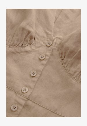 Beige linane topp, millel on kogutud rinnaosa, viis nööpi, millest igaühel on neli auku, ning peened õmblusdetailid. Pehme tekstuur ja kerge.