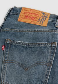 Jeans de mezclilla azul oscuro con una patch de cuero marrón que presenta el logo de Levi Strauss. Bolsillo trasero con costuras naranjas y una pequeña etiqueta roja.