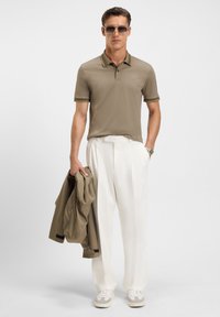 Oliven polo skjorte med tre knappelukkinger og kontrastkrage, kombinert med hvite plissert bukser og beige sneakers. Holder en matchende jakke.