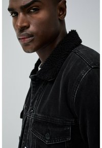 Chaqueta de mezclilla negra con un cuello de pelaje negro, costuras prominentes y un detalle de bolsillo en el frente. La tela parece ligeramente desgastada.