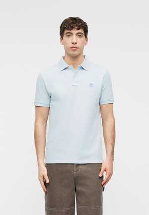 Mladý muž s kučeravými vlasmi, ktorý má na sebe svetlomodré polo tričko s krátkym rukávom a tmavohnedé nohavice, stojaci na bielom jednoduchom pozadí.