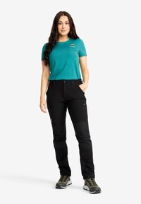 Kvinna stående med en hand i fickan på svarta cargobyxor, iförd en teal kortärmad skjorta och olivgröna vandringsskor på vit bakgrund.