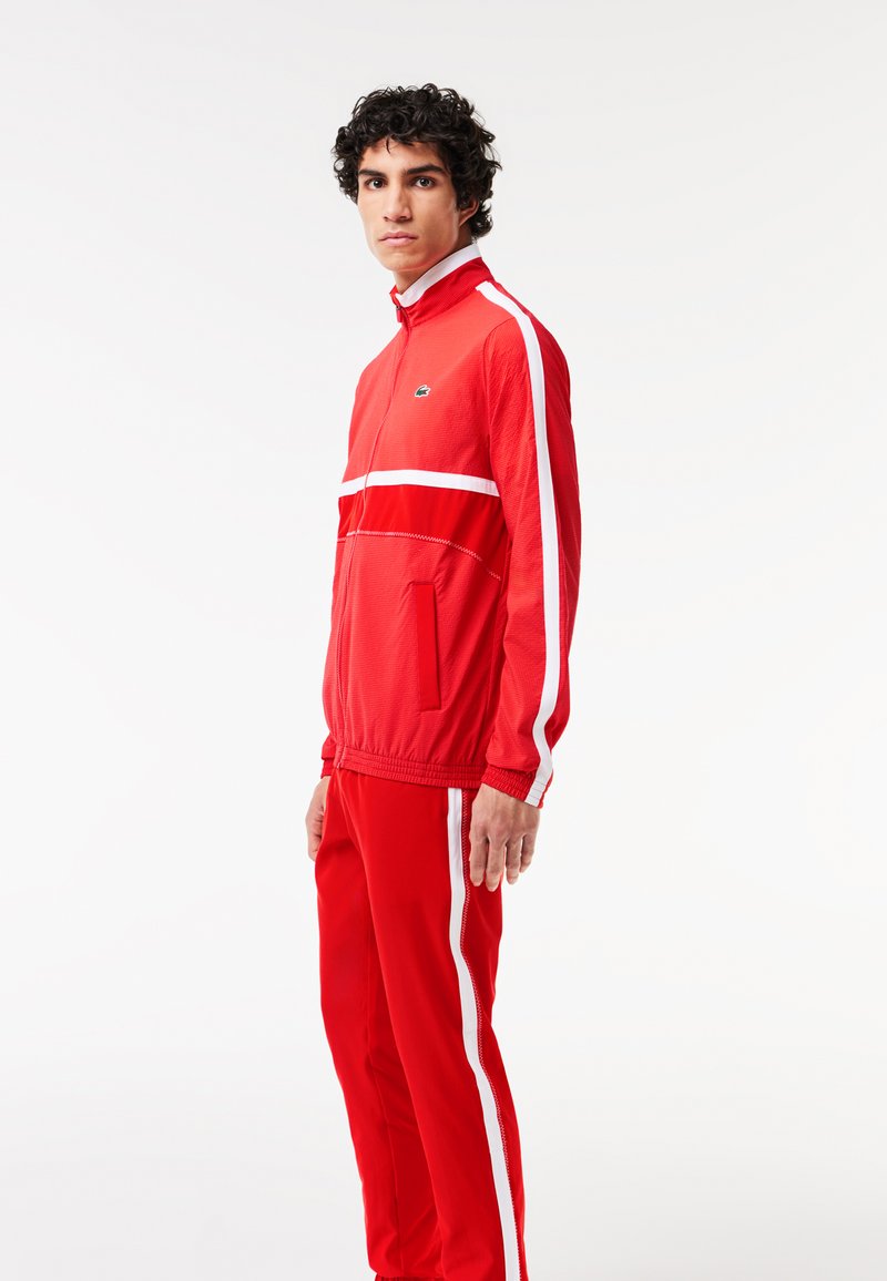 Ensemble de survêtement rouge en tissu léger. La veste présente des rayures blanches, une fermeture éclair et une poche. Le pantalon a des rayures assorties et une taille élastique.