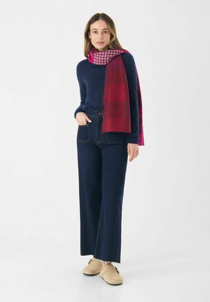 Pull en tricot bleu marine, jeans taille haute à large jambe, chaussures beige à enfiler, et une écharpe à carreaux rose drapée autour du cou.