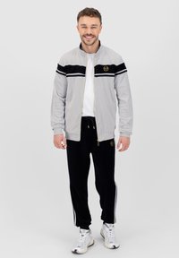 Chaqueta gris con cierre y rayas negras, acompañada de joggers negros con rayas laterales blancas. Se lleva con zapatillas blancas.
