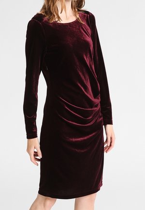 Kobieta w długiej do kolan, ciemnoczerwonej welurowej sukience z marszczeniami z przodu, na białym tle.