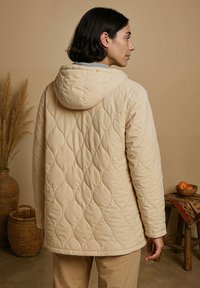 Beige gewatteerde jas met een capuchon, voorzien van een golvend patroon en een ronde zoom. De binnenkant toont een grijze voering en het model is van achteren gezien.