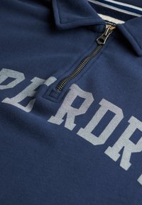 Granatowa koszulka polo z zamkiem w kołnierzu, z dużym jasnoszarym napisem. Materiał wydaje się miękki i teksturowany, odpowiedni do noszenia na co dzień.