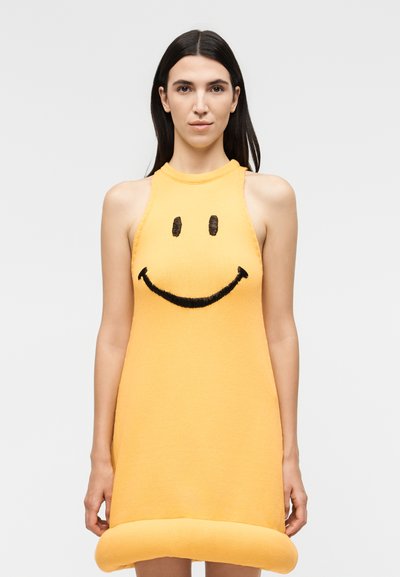Femme aux longs cheveux foncés portant une robe jaune sans manches avec un grand visage souriant noir sur le devant, sur un fond uni.