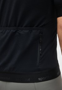 Κοντινό πλάνο της πίσω τσέπης μαύρης υφασμάτινης φανέλας ποδηλασίας με κάθετο φερμουάρ, φορεμένη πάνω από γκρι παντελόνι, με εμφανές μέρος του χεριού ενός ατόμου.