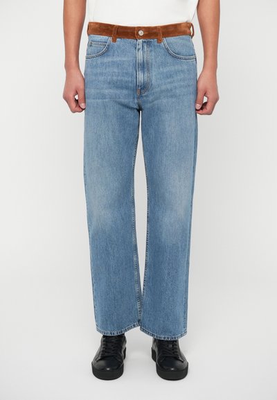 Jeans a gamba larga blu chiaro in denim, con una cintura in suede marrone, cinque tasche e orlo cucito, abbinati a sneakers nere.