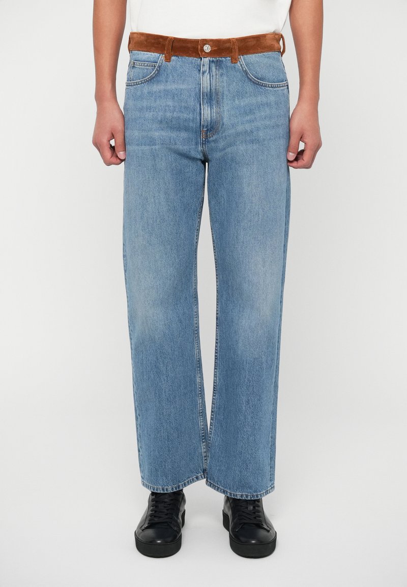 Hellblaue weit geschnittene Jeans aus Denim, ausgestattet mit einem braunen Wildledergürtel, fünf Taschen und einem gestickten Saum, kombiniert mit schwarzen Sneakers.