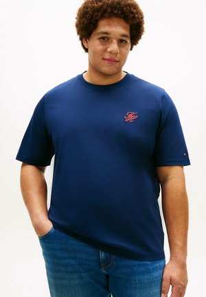 Uomo con capelli ricci e lentiggini, che indossa una maglietta blu navy con un piccolo logo rosso e jeans blu, in piedi con una mano in tasca.