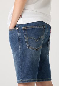 Denim shorts in medium wash blue, featuring two back pockets with distinctive stitching, and slightly frayed hem. Cotton fabric with a smooth texture.