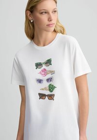 T-shirt en coton blanc avec cinq paires de lunettes de soleil graphiques dans différentes couleurs et motifs, incluant des designs verts, roses, violets et bruns.
