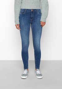 Jeans de mezclilla azul de corte ajustado con una textura suave, cinco bolsillos y cierre de botón metálico. Combinados con zapatillas grises y un suéter de punto.