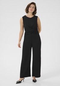 Jumpsuit nero senza maniche con dettaglio frontale attorcigliato, gambe ampie e texture morbida, abbinato a tacchi a punta neri.