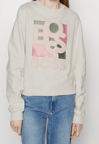 Felpa beige in cotone con un grande logo "BOSS" multicolore sul davanti. Vestibilità comoda con polsini e orlo a costine.