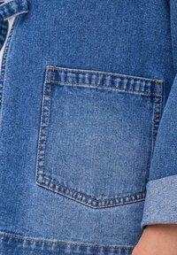 Zapnjen denim jakne s poudarjeno modro barvo, žepom na prsih s ravnim rezom ter kontrastnim šivom ob robovih in manžetah rokavov.