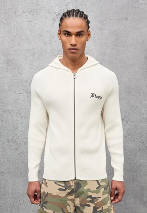 Junger Mann mit geflochtenen Haaren, der einen weißen Hoodie mit Reißverschluss und dem Schriftzug "Utopia" sowie eine Camouflage-Hose trägt, vor einer grauen Betonwand.