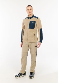 Mladý muž oblečený v béžové fleecové bundě s námořnickými doplňky, béžových cargo kalhotách a modro-černých turistických botách, stojící na bílém pozadí.