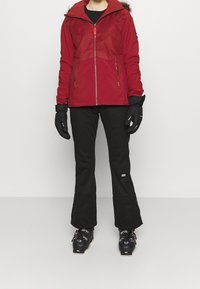 Osoba w czerwonej zimowej kurtce, czarnych spodniach narciarskich, czarnych rękawiczkach i czarnych butach narciarskich, stojąca na jasnym, jednolitym tle.