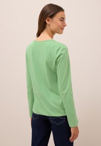 Frau mit braunen Haaren, die seitwärts blickt, trägt ein hellgrünes Langarmshirt und dunkelblaue Jeans vor einem einfarbigen Hintergrund.