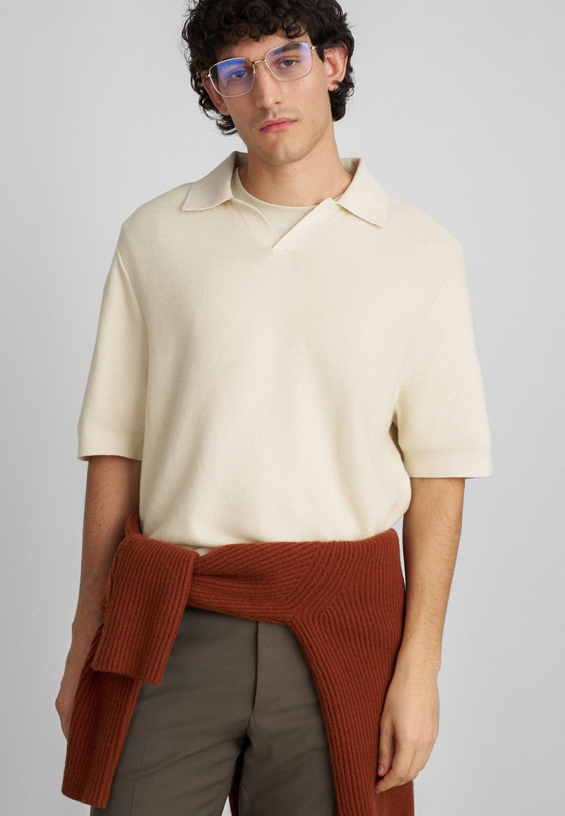 Krema kratka majica s kratkimi rokavi in V-izrezom, z teksturirano površino. Rjave rebraste pletenine okoli pasu, ki dopolnjuje bež hlače.