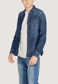 Camicia di jeans di colore blu scuro con chiusura a bottoni, due tasche sul petto, maniche lunghe e colletto classico. Abbinata a jeans blu chiaro.