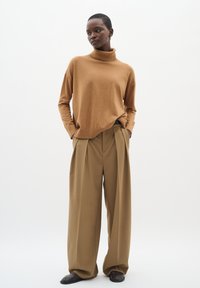 Brun rullekravesweater i blødt materiale, kombineret med beige bukser med vidde og plisseringer. Modellen står med hænderne i lommerne.