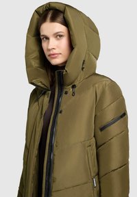 Kvinna iklädd en olivgrön pufferjacka med stor huva och svarta dragkedjedetaljer, vänd något åt vänster mot en enfärgad bakgrund.