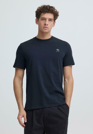 Ung mann med krøllete hår iført en enkel svart kortermet t-skjorte med en liten hvit logo på venstre bryst, stående foran en lys bakgrunn.