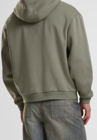 Felpa con cappuccio verde oliva con tasca a marsupio e polsini a coste, abbinata a jeans in denim chiaro con dettagli sulle tasche posteriori.