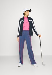 Outfit da golf caratterizzato da una maglia polo rosa, giacca blu navy con strisce bianche, pantaloni a righe blu e un cappellino bianco. Bastone tenuto nella mano destra.