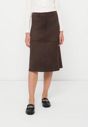 Donna che indossa una gonna marrone scuro fino al ginocchio abbinata a scarpe slip-on nere con dettagli in catena dorata e una maglia bianca a maniche lunghe.