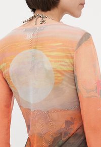 Top transparente de manga larga con un gráfico de puesta de sol en tonos naranjas y rosas, con un cierre de lazo en la espalda y un acento de patrón de cuadros.