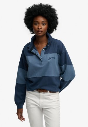 Frau mit lockigem Haar, die einen zweifarbigen blauen Superdry-Pullover und weiße Hosen mit einem braunen geflochtenen Gürtel trägt, steht vor einem weißen Hintergrund.