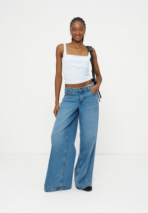 Femme debout portant un haut blanc sans manches, un jean bleu à jambes larges, des chaussures noires et un sac à bandoulière noir sur un fond blanc.