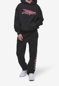 Svart huvtröja med en rosa grafisk logo och ficka fram, kombinerad med matchande svarta mjukisbyxor med vit logotryck.