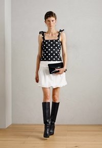 Top en dentelle noire et blanche à pois avec des bretelles en nœud, associé à une jupe blanche et des bottes noires au-dessus du genou, tenant une pochette noire.