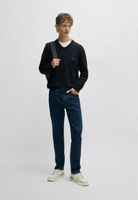 Svart v-ringad tröja, avslappnade denimjeans i mörkblå färg, och beige sneakers. Modellen håller en grå remväskan med en liten logotyp-patch.