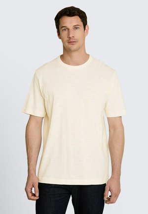 Man met kort donker haar die een effen lichtbeige T-shirt met korte mouwen en donkere broek draagt, staand tegen een witte achtergrond.