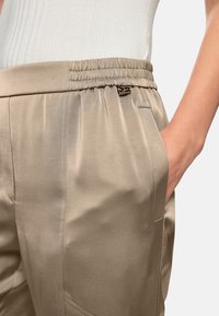 Beige satin trousers with an elastic waistband, side pockets, and subtle pleats. The fabric has a soft sheen and smooth texture.