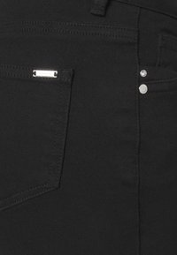 Pantalones de mezclilla negros que presentan un bolsillo estándar con una placa de logo plateada, dos remaches metálicos y costuras visibles.