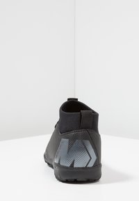 Mustat urheilukengät, joissa on teksturoitu pinta, korkea neulekaulus ja heijastava logo takana. Kumia oleva pohja tarjoaa hyvän pidon.