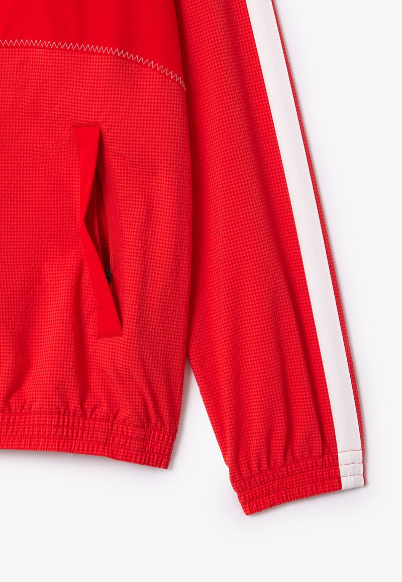 Veste pullover rouge en tissu texturé, avec une poche zippée, des poignets élastiques et une bande latérale blanche pour le contraste.